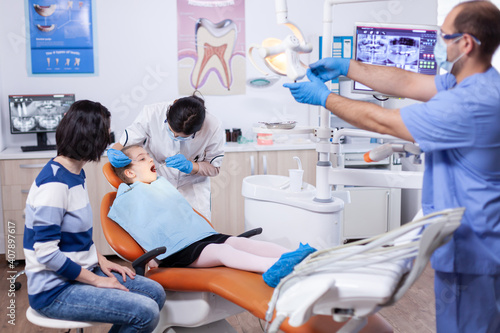 Kid patient in dental office getting teeth treatment sitting on dental chair wearing bib with mouth open. Dentistry specialist during child cavity consultation in stomatology office using modern