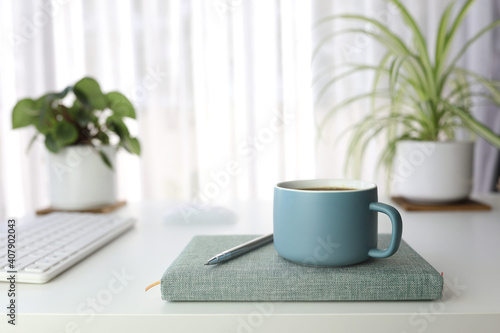 Blue modern matte coffee cup and pencil and green notebook on white table with plants pot and keyboard