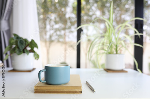 Blue modern matte coffee cup and pencil and brown notebook on white table with plants pot and keyboard