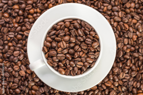 A white coffee cup full of fresh coffee beans on roasted coffee beans background top view