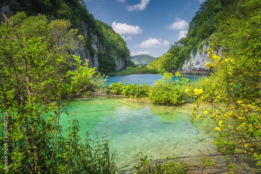 Fototapeta premium Tourists walking on footpath under tall cliff in a valley with lakes, framed by branches. Plitvice Lakes National Park UNESCO World Heritage, Croatia