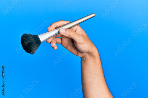 Hand of hispanic man holding makeup brush over isolated blue background.
