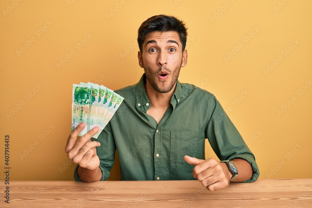 Young handsome man holding 50 hong kong dollars banknotes scared and amazed with open mouth for surprise, disbelief face Young handsome man holding 50 hong kong dollars banknotes scared and amazed with open mouth for surprise, disbelief face