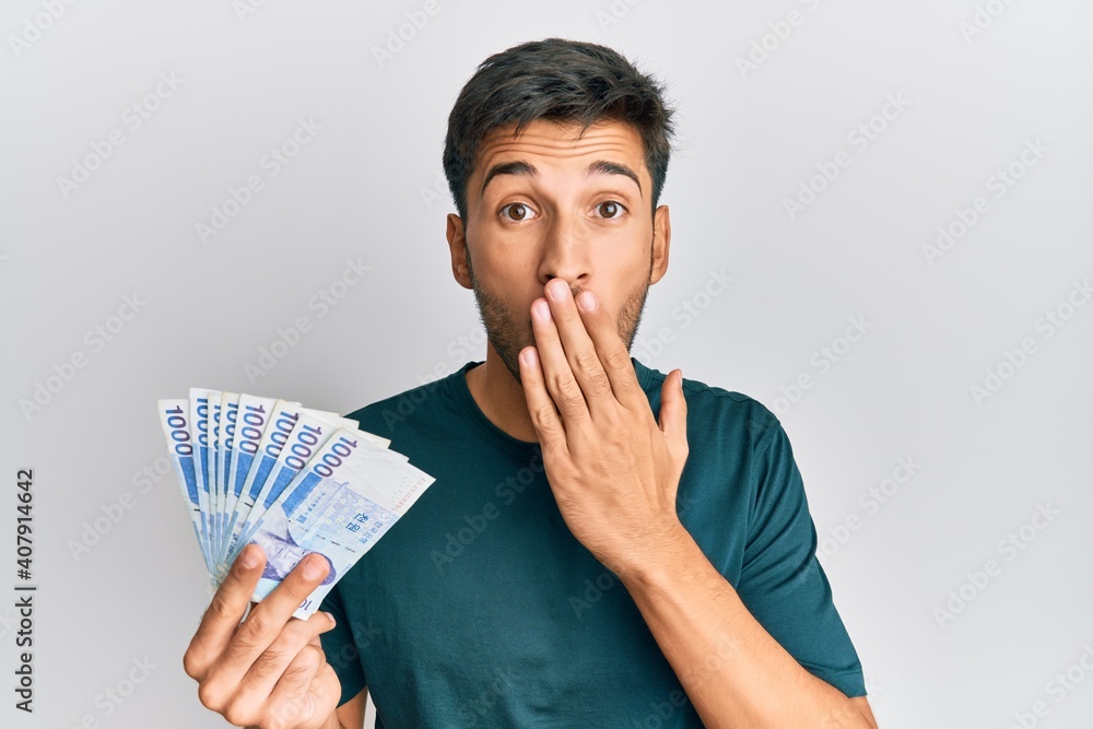 Young handsome man holding 1000 south korean won banknotes covering mouth with hand, shocked and afraid for mistake. surprised expression Young handsome man holding 1000 south korean won banknotes covering mouth with hand, shocked and afraid for mistake. surprised expression