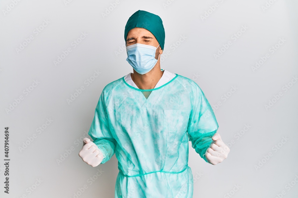 Young handsome man wearing surgeon uniform and medical mask excited for success with arms raised and eyes closed celebrating victory smiling. winner concept. Young handsome man wearing surgeon uniform and medical mask excited for success with arms raised and eyes closed celebrating victory smiling. winner concept.