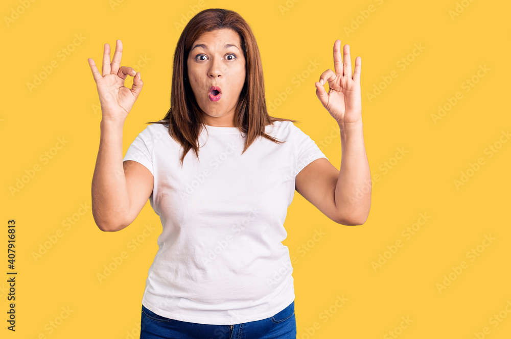 Middle age latin woman wearing casual white tshirt looking surprised and shocked doing ok approval symbol with fingers. crazy expression Middle age latin woman wearing casual white tshirt looking surprised and shocked doing ok approval symbol with fingers. crazy expression