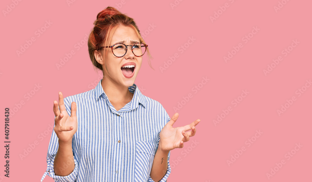 Young redhead woman wearing casual clothes and glasses crazy and mad shouting and yelling with aggressive expression and arms raised. frustration concept. Young redhead woman wearing casual clothes and glasses crazy and mad shouting and yelling with aggressive expression and arms raised. frustration concept.