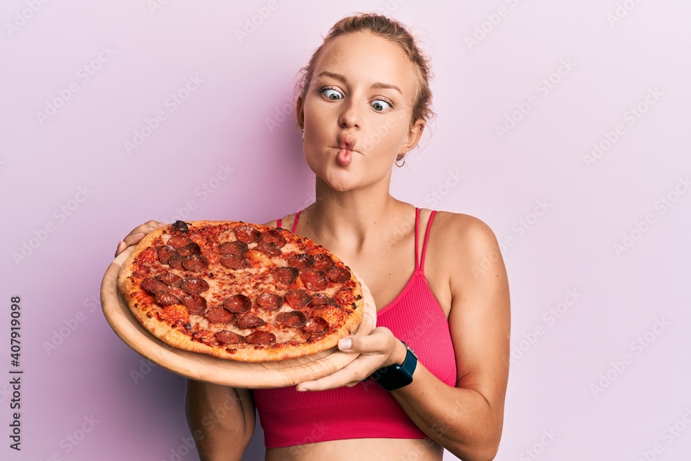 Beautiful caucasian woman holding italian pizza making fish face with mouth and squinting eyes, crazy and comical. Beautiful caucasian woman holding italian pizza making fish face with mouth and squinting eyes, crazy and comical.