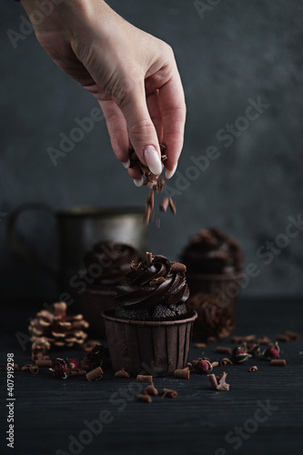 Several muffins or cupcakes with chocolate shaped cream at black table. Festive candle burns on a chocolate cake. A woman's hand crumbles grated chocolate onto a cake
