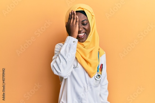 Beautiful african young woman wearing doctor uniform and hijab covering one eye with hand, confident smile on face and surprise emotion.