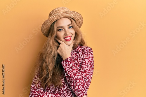 Beautiful blonde caucasian woman wearing summer hat looking confident at the camera smiling with crossed arms and hand raised on chin. thinking positive.