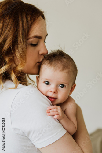 Beautiful mom hugs and kisses the newborn in the studio on a white background. A young mother in a white bodysuit hugs and kisses her newborn daughter in the studio.