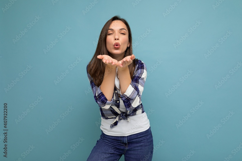 Young positive happy beautiful winsom brunette woman with sincere emotions wearing check shirt poising isolated over blue background with copy space and giving kiss Young positive happy beautiful winsom brunette woman with sincere emotions wearing check shirt poising isolated over blue background with copy space and giving kiss