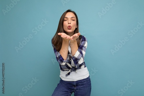 Young positive happy beautiful winsom brunette woman with sincere emotions wearing check shirt poising isolated over blue background with copy space and giving kiss © Ivan Traimak