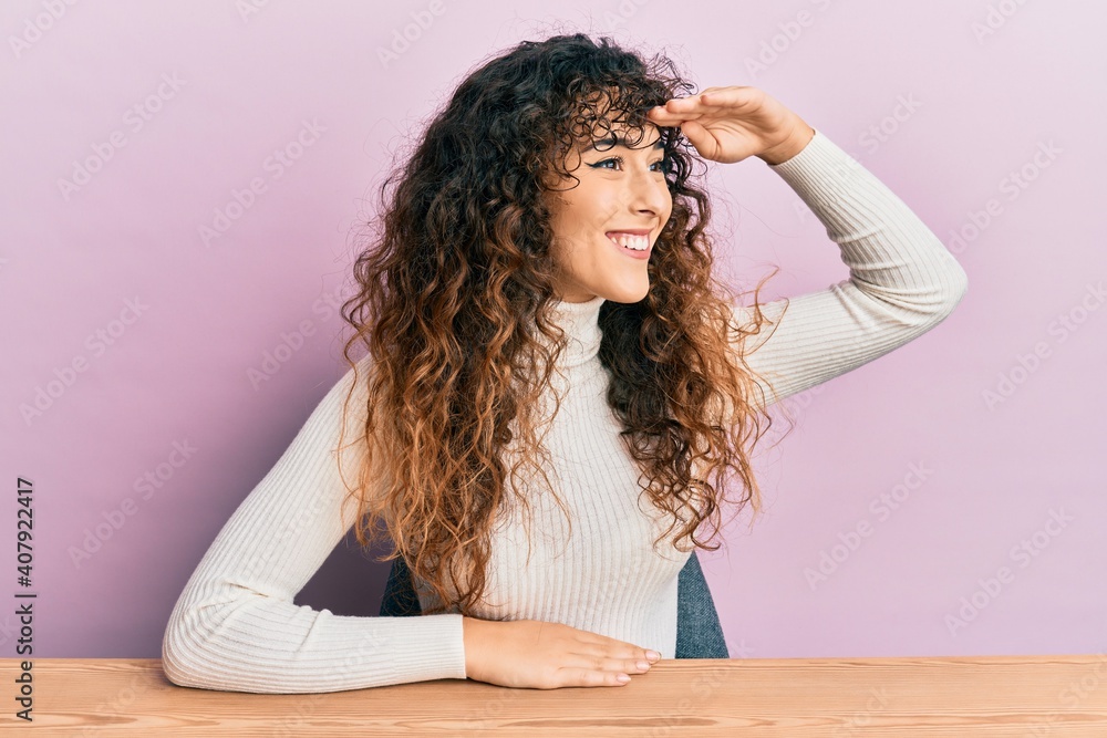 Young hispanic girl wearing casual clothes sitting on the table very happy and smiling looking far away with hand over head. searching concept.