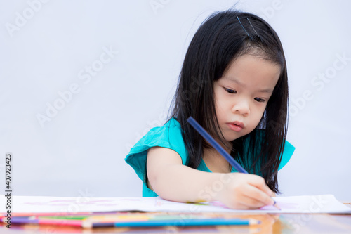 Asian adorable girl is doing homework. Children paint with wooden colors on a coloring book. White paper. Child learn art. On a white wall background. Kid is 4 years old. Home school. Stay at home. photo