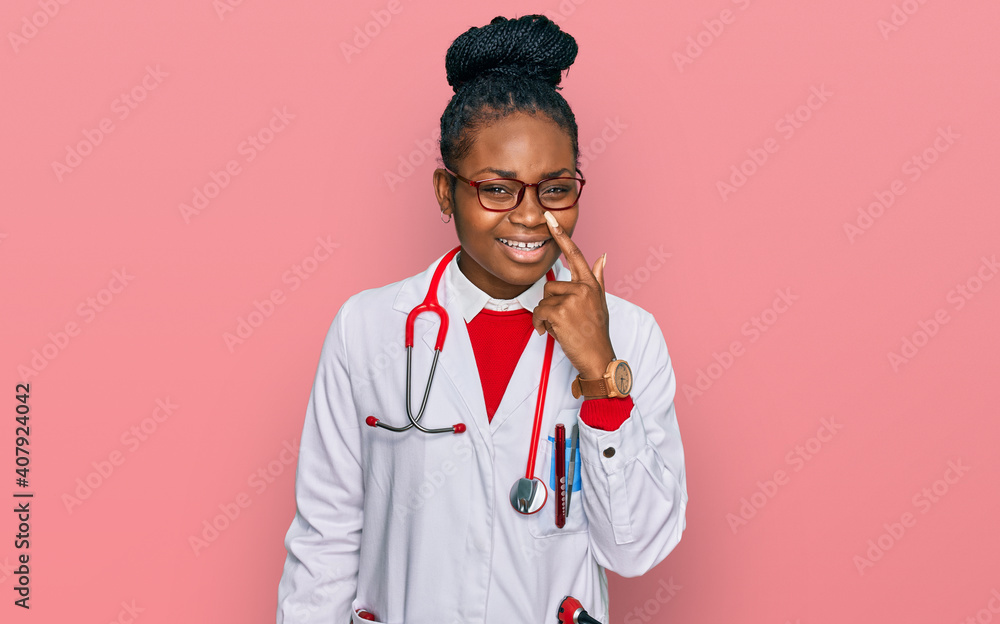 Young african american woman wearing doctor uniform and stethoscope pointing with hand finger to face and nose, smiling cheerful. beauty concept Young african american woman wearing doctor uniform and stethoscope pointing with hand finger to face and nose, smiling cheerful. beauty concept