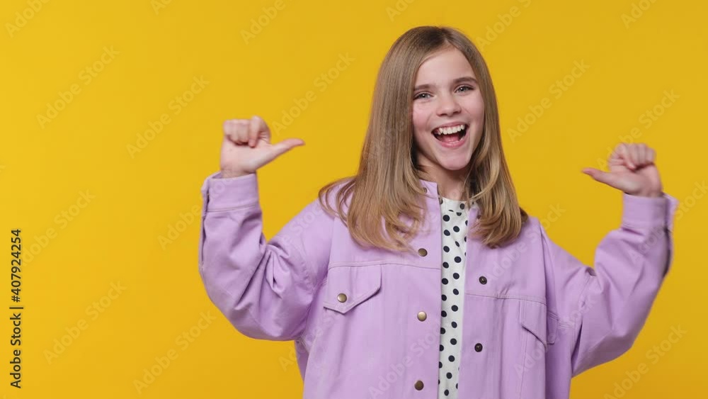 Cheerful excited little blonde kid girl 12-13 years old wearing violet casual clothes isolated on bright yellow color wall background studio. Childhood lifestyle concept. Pointing thumbs on herself Cheerful excited little blonde kid girl 12-13 years old wearing violet casual clothes isolated on bright yellow color wall background studio. Childhood lifestyle concept. Pointing thumbs on herself