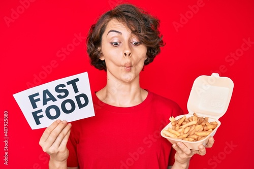 Young hispanic woman holding potato chip and fast food banner making fish face with mouth and squinting eyes  crazy and comical.