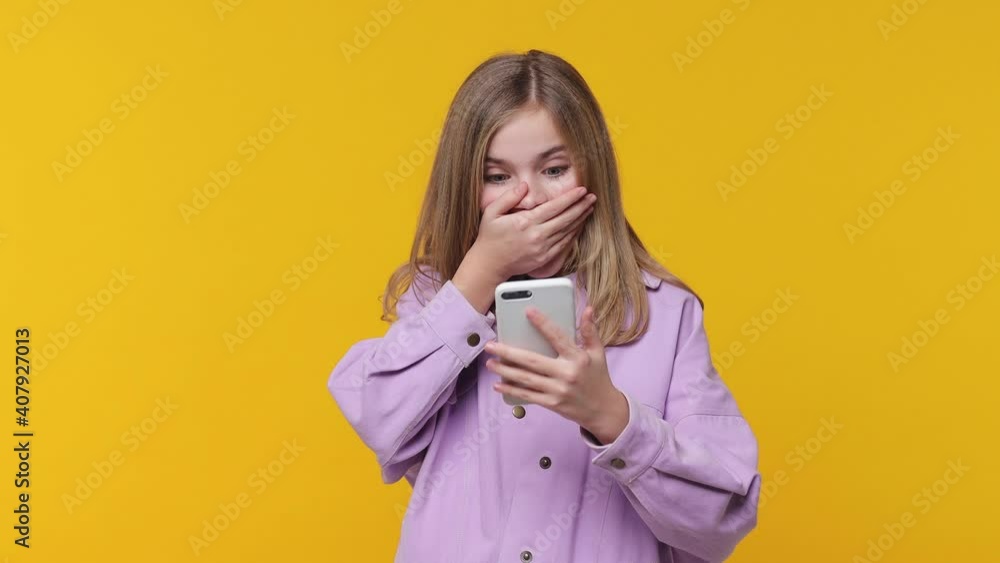 Surprised little blonde kid girl 12-13 in violet clothes isolated on yellow background studio. Childhood lifestyle concept. Using mobile cell phone browsing say wow cover mouth doing winner gesture Surprised little blonde kid girl 12-13 in violet clothes isolated on yellow background studio. Childhood lifestyle concept. Using mobile cell phone browsing say wow cover mouth doing winner gesture