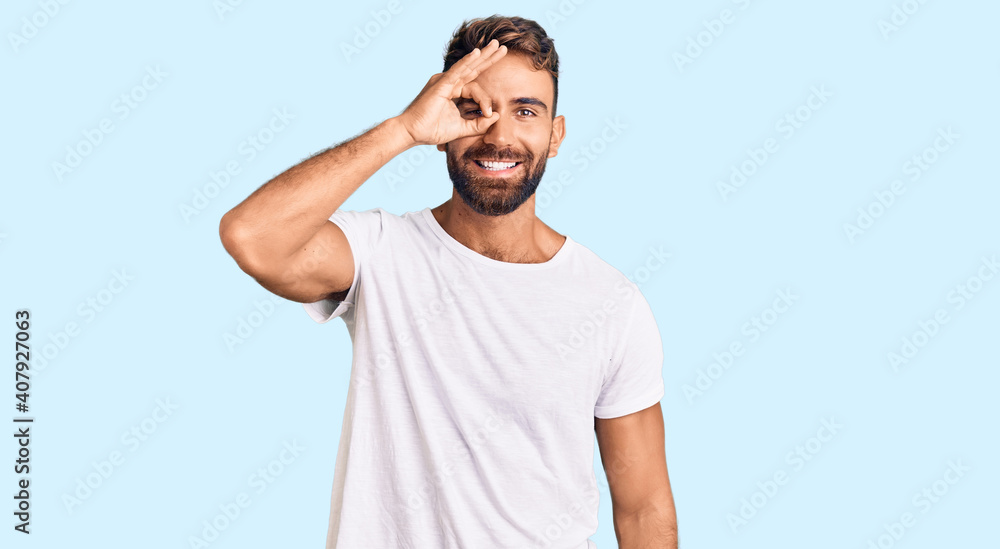 Young hispanic man wearing casual white tshirt doing ok gesture with hand smiling, eye looking through fingers with happy face. Young hispanic man wearing casual white tshirt doing ok gesture with hand smiling, eye looking through fingers with happy face.