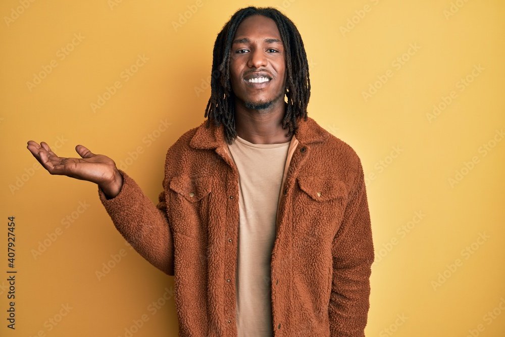 African american man with braids wearing brown retro jacket smiling cheerful presenting and pointing with palm of hand looking at the camera. African american man with braids wearing brown retro jacket smiling cheerful presenting and pointing with palm of hand looking at the camera.