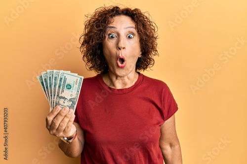 Beautiful middle age mature woman holding 20 dollars banknotes scared and amazed with open mouth for surprise, disbelief face