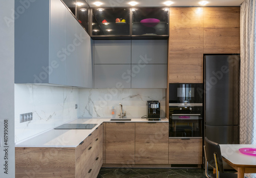 The interior of the studio kitchen is decorated in gray tones with wooden veneers. Kitchen appliances are black. The table top is white.