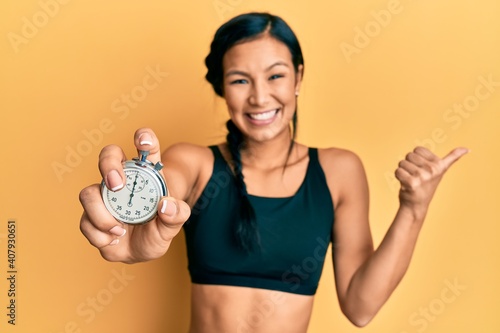 Beautiful hispanic woman wearing sportswear holding stopwatch pointing thumb up to the side smiling happy with open mouth