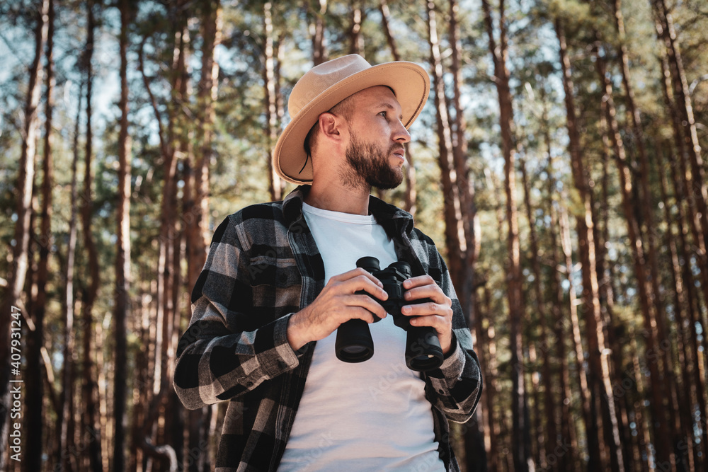 Man tourist in a hat and gray plaid shirt looks through binoculars