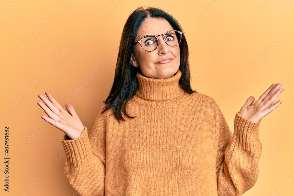 Middle age brunette woman wearing casual winter sweater over yellow background clueless and confused with open arms, no idea and doubtful face. Middle age brunette woman wearing casual winter sweater over yellow background clueless and confused with open arms, no idea and doubtful face.