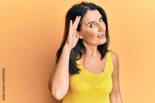 Middle age brunette woman wearing casual clothes over yellow background smiling with hand over ear listening and hearing to rumor or gossip. deafness concept.