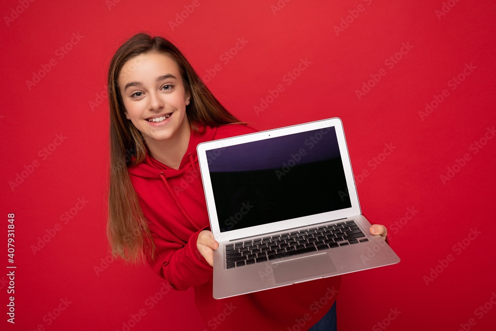 Close-up portrait photo of beautiful happy smiling girl with long hair wearing red hoodie holding computer laptop looking at camera isolated over red wall background Close-up portrait photo of beautiful happy smiling girl with long hair wearing red hoodie holding computer laptop looking at camera isolated over red wall background