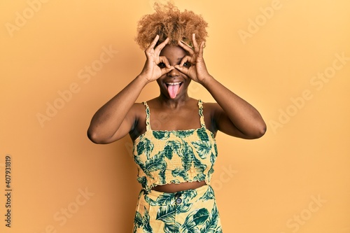 Young african woman with afro hair wearing summer dress doing ok gesture like binoculars sticking tongue out  eyes looking through fingers. crazy expression.