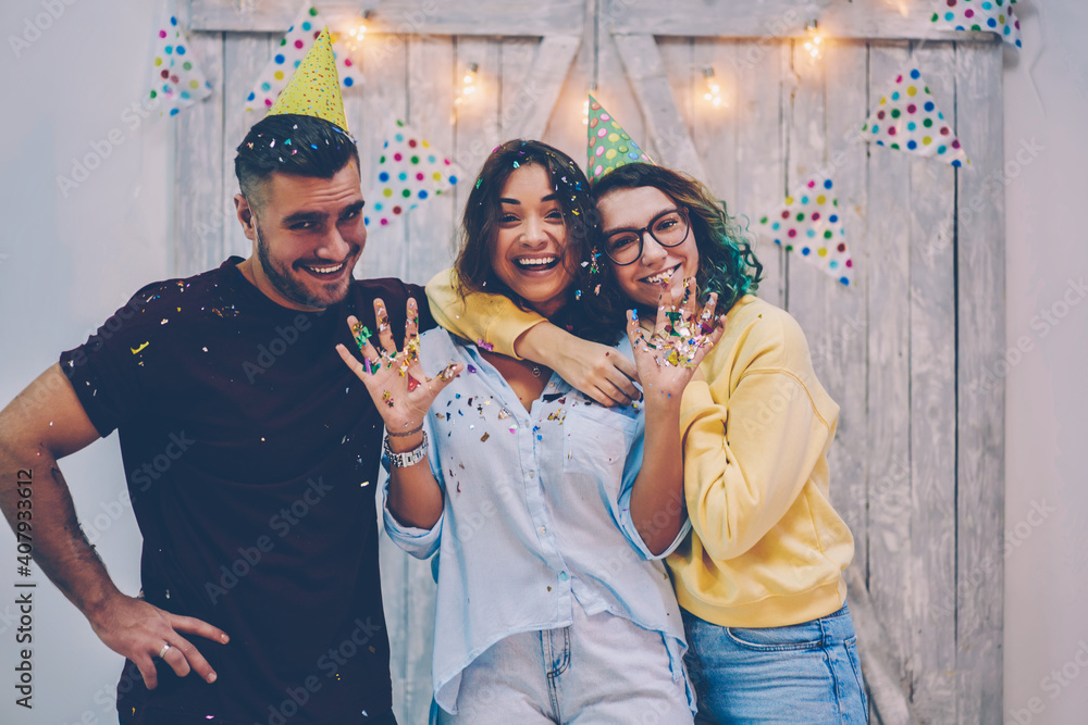 Obraz premium Portrait of happy young hipster guys in festive cone hats celebrating birthday together, group of overjoyed male and female guests of anniversary party hugging and looking at camera with decorations
