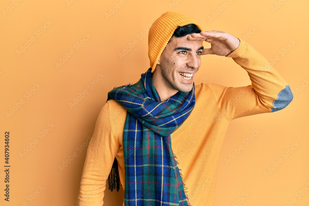 Handsome hispanic man wearing wool hat and winter scarf very happy and smiling looking far away with hand over head. searching concept. Handsome hispanic man wearing wool hat and winter scarf very happy and smiling looking far away with hand over head. searching concept.