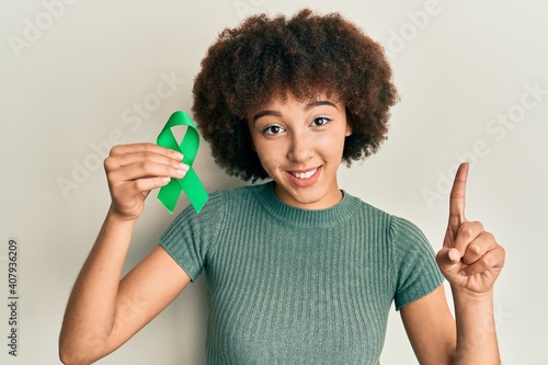 Young hispanic girl holding support green ribbon surprised with an idea or question pointing finger with happy face, number one