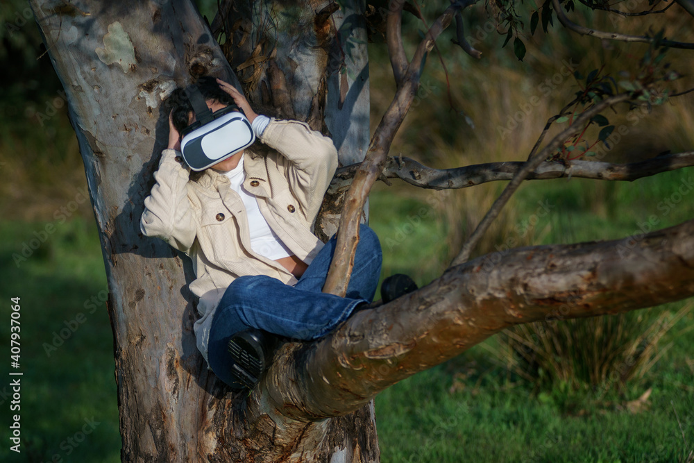 Naklejka premium Vista de frente de una joven hispana de cabello negro y rizado que lleva un casco de realidad virtual y disfruta de la experiencia subida en un árbol en el bosque vestida de manera casual en invierno.