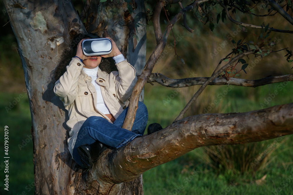 Naklejka premium Vista de frente de una joven hispana de cabello negro y rizado que lleva un casco de realidad virtual y disfruta de la experiencia subida en un árbol en el bosque vestida de manera casual en invierno.