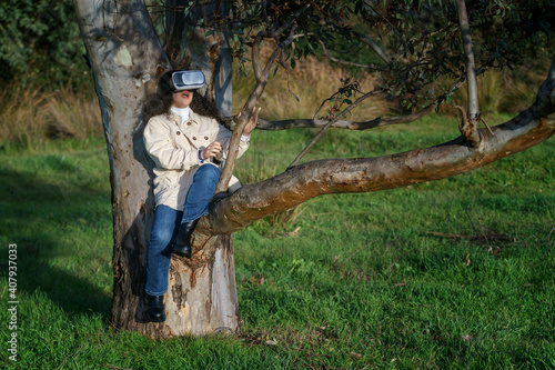 Vista de frente de una joven hispana de cabello negro y rizado que lleva un casco de realidad virtual y disfruta de la experiencia subida en un árbol en el bosque vestida de manera casual en invierno.