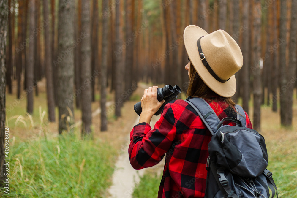 Young woman tourist with backpack, hat and red plaid shirt and looks through binoculars in the forest