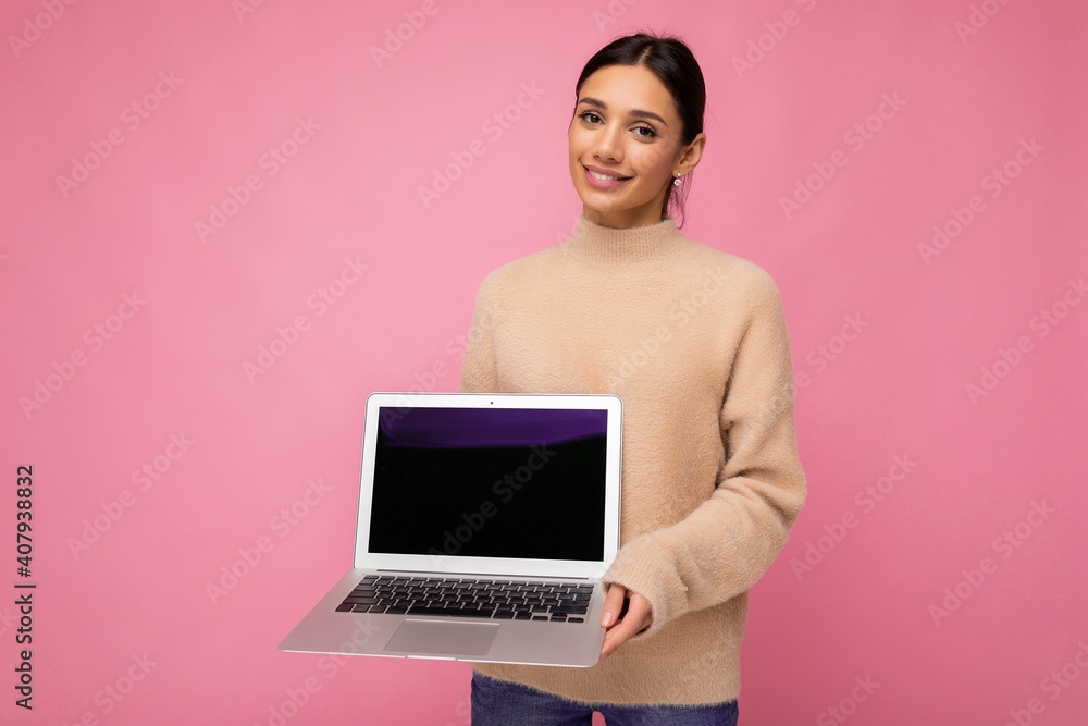 beautiful smiling young woman with dark hair looking at camera holding computer laptop with empty monitor screen with mock up and copy space wearing light colour sweater isolated on pink wall beautiful smiling young woman with dark hair looking at camera holding computer laptop with empty monitor screen with mock up and copy space wearing light colour sweater isolated on pink wall