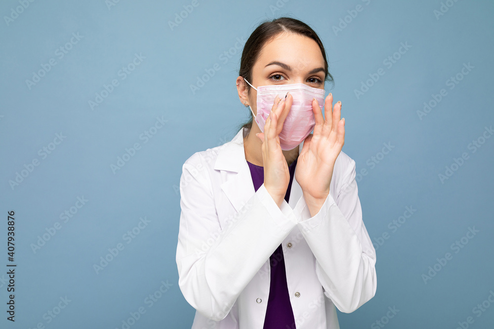 Young beautiful woman in reusable virus protective mask on face against coronavirus whispering secret isolated on the blue background wall Young beautiful woman in reusable virus protective mask on face against coronavirus whispering secret isolated on the blue background wall