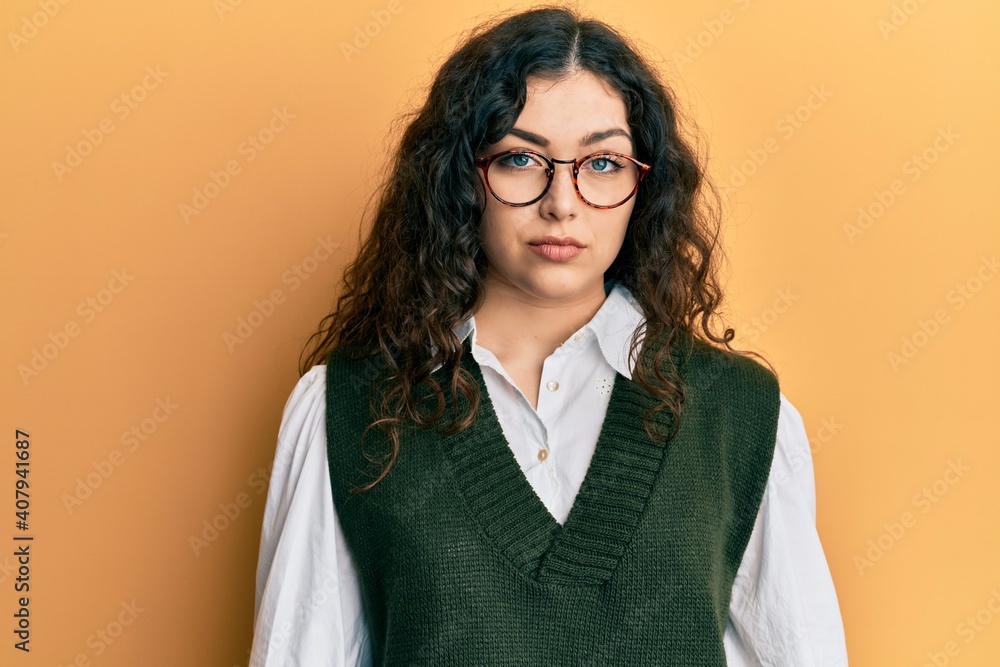 Young brunette woman with curly hair wearing casual clothes and glasses with serious expression on face. simple and natural looking at the camera.