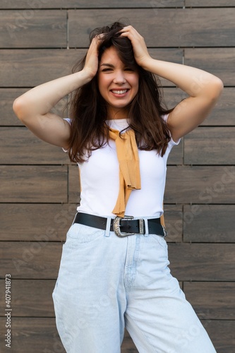 Vertical ptoto of young beautiful positive happy brunette woman standing against brown wall in the street and wearing stylish outfit photo