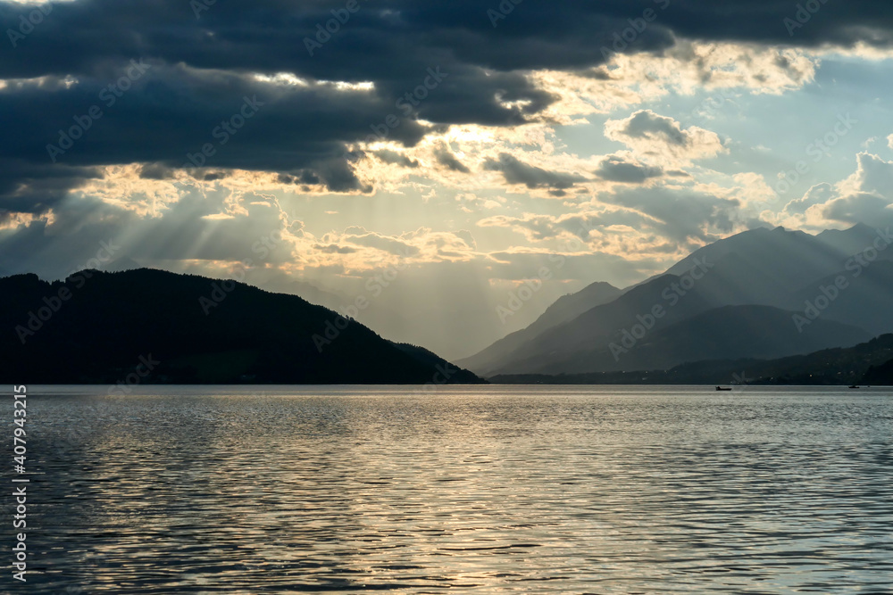 A sunset by Millstaetter lake in Austria. The lake is surrounded by high Alps. Calm surface of the lake reflecting the sunbeams. The sun sets behind the mountains. Thick clouds above. Natural beauty A sunset by Millstaetter lake in Austria. The lake is surrounded by high Alps. Calm surface of the lake reflecting the sunbeams. The sun sets behind the mountains. Thick clouds above. Natural beauty