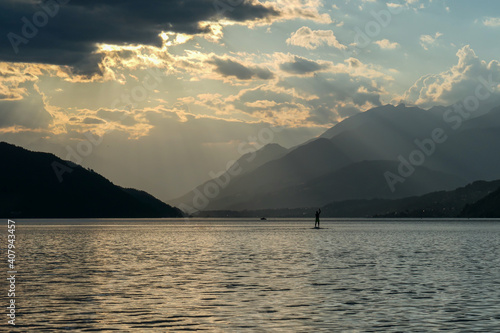 A man stand up paddling during the sunset on Millstaetter lake in Austria. The lake is surrounded by high Alps. Calm surface of the lake reflecting the sunbeams. The sun sets behind the mountains.