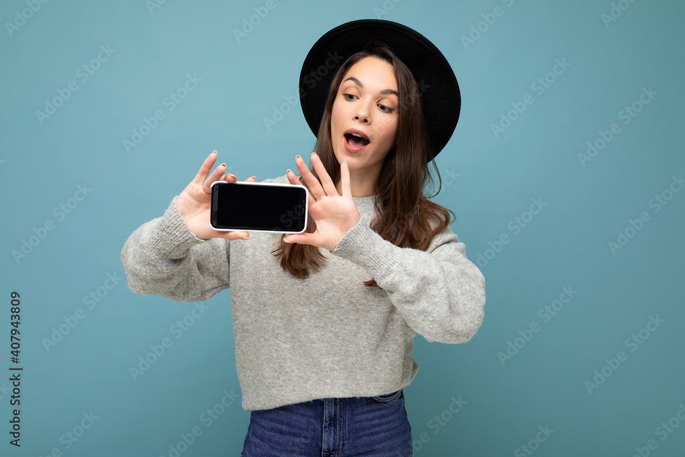 Attractive young smiling woman wearing black hat and grey sweater holding smartphone looking down isolated on background Attractive young smiling woman wearing black hat and grey sweater holding smartphone looking down isolated on background