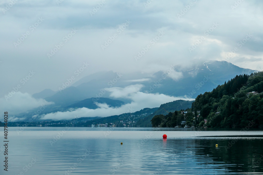 A panoramic view on the Millstaettersee lake from its shore. The lake is surrounded by high mountains. A few red buoys floating on the surface. Thick and dark clouds above. Serenity and calmness