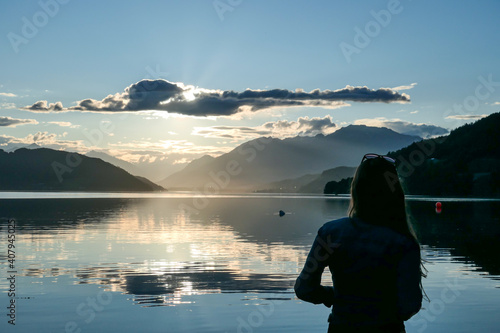 A woman standing at the shore of Millstaetter lake during the sunset. High Alps around. The sun sets behind thick clouds. Calm surface of the lake reflects the orange sky and the mountains. Day end photo